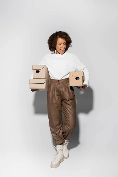 Pleine longueur de femme afro-américaine étonnée avec des cheveux bouclés tenant boîtes de livraison sur gris — Photo de stock