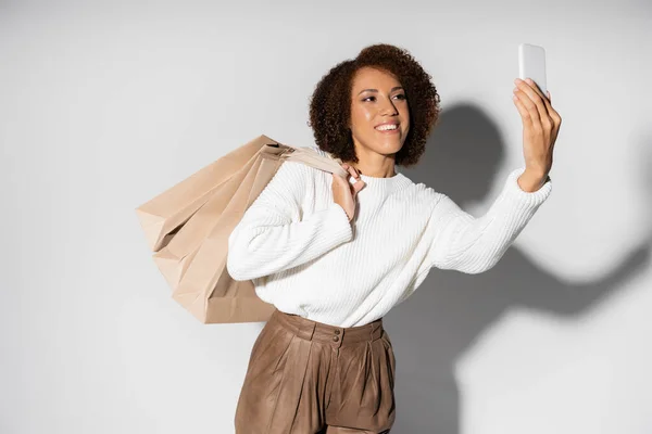 Улыбающаяся африканская американка в осеннем наряде держит сумки с покупками и делает селфи на серой — стоковое фото