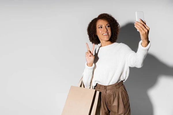 Heureuse femme afro-américaine en tenue automnale tenant des sacs à provisions et montrant la paix tout en prenant selfie sur gris — Photo de stock