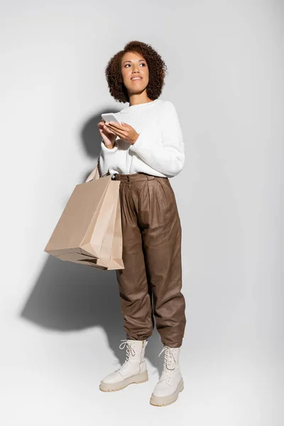 Pleine longueur de femme afro-américaine heureuse en tenue automnale tenant des sacs à provisions et utilisant un smartphone sur gris — Photo de stock