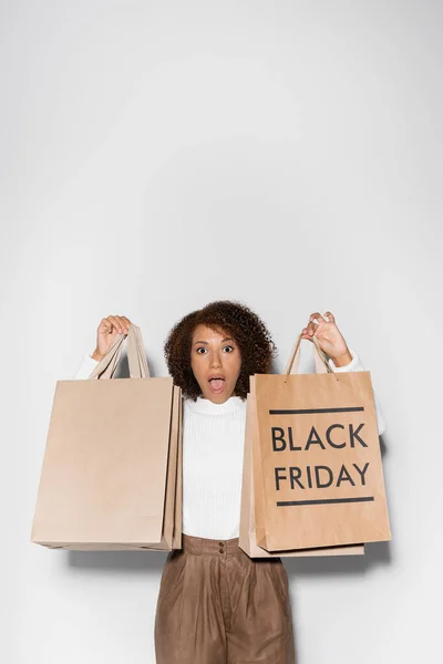 Femme afro-américaine choquée en tenue automnale tenant des sacs à provisions avec lettrage noir vendredi sur gris — Photo de stock