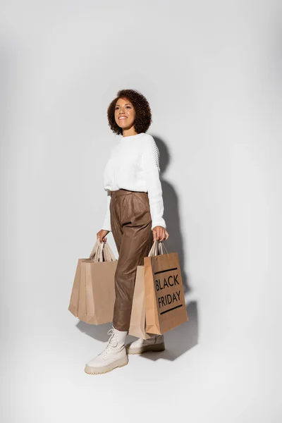 Pleased african american woman in autumnal outfit holding shopping bags with black friday lettering on grey — Stock Photo