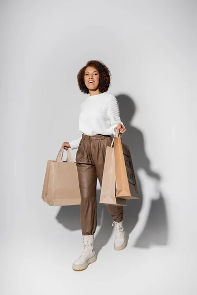 Счастливая африканская американка в осеннем наряде держит сумки с покупками и ходит по серой — стоковое фото