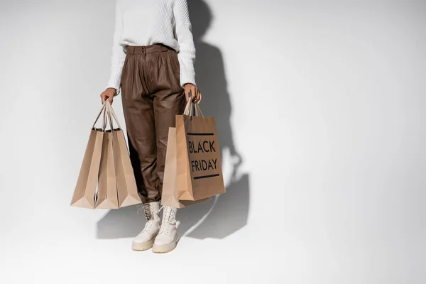 Vista parcial de mujer afroamericana en traje otoñal sosteniendo bolsas de compras con letras de viernes negro en gris - foto de stock