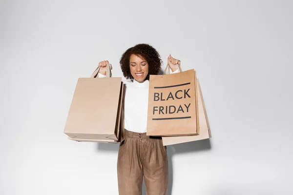 Deliziata donna afro-americana con gli occhi chiusi e capelli ricci che tengono borse della spesa con scritte venerdì nero su grigio — Foto stock