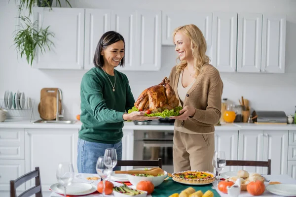 Cheerful interracial mother and daughter holding thanksgiving turkey near table at home — Stock Photo