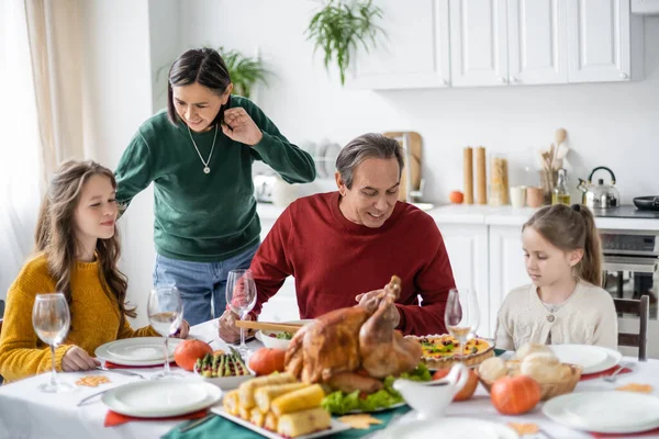 Abuelos multiétnicos sonrientes hablando con niños cerca de la cena de Acción de Gracias en casa - foto de stock