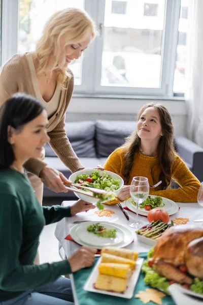 Lächelnde Tochter schaut Mutter mit Salat in der Nähe verschwommener multiethnischer Großeltern zu Hause an — Stockfoto