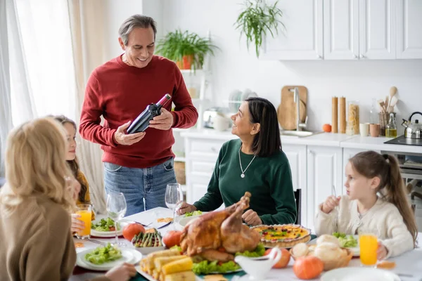 Старший мужчина держит вино возле многонациональной семьи и ужин в честь Дня благодарения дома — стоковое фото
