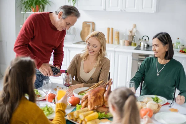 Senior man pouring wine near smiling multicultural family during thanksgiving dinner at home — Stock Photo