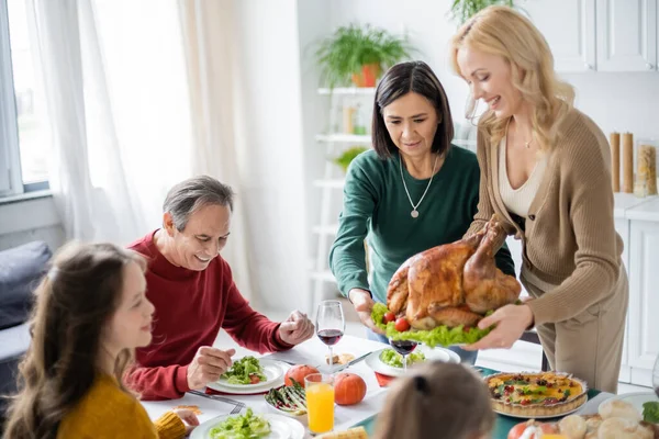 Femmes multiethniques tenant la dinde près d'une famille souriante et dîner d'action de grâces à la maison — Photo de stock