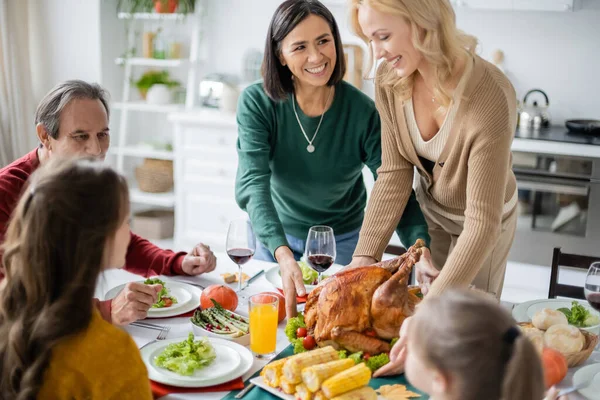 Mãe multiétnica olhando para a filha segurando peru perto da família turva e jantar de ação de graças em casa — Fotografia de Stock