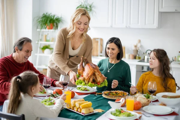 Mujer positiva sosteniendo pavo cerca de la familia multicultural y comida en casa - foto de stock