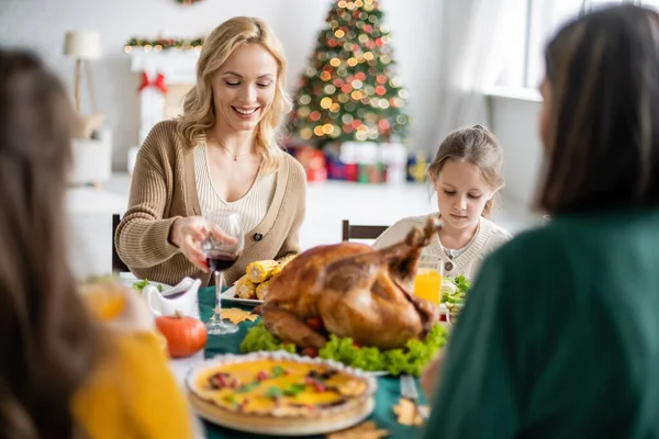 Donna sorridente che tiene un bicchiere di vino vicino alla famiglia e cena di ringraziamento offuscata a casa — Foto stock