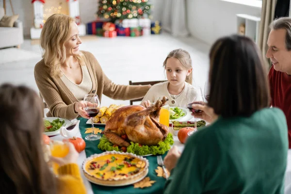 Madre sonriente mirando al niño cerca de la familia borrosa y la cena de acción de gracias en casa - foto de stock