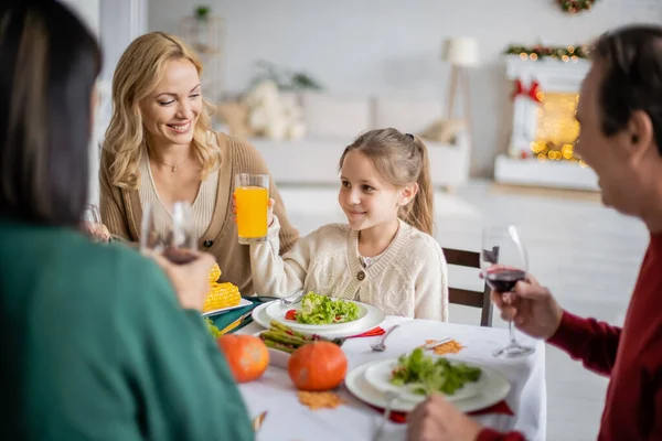 Smiling girl holding orange juice near mother and blurred grandparents during thanksgiving celebration — Stock Photo