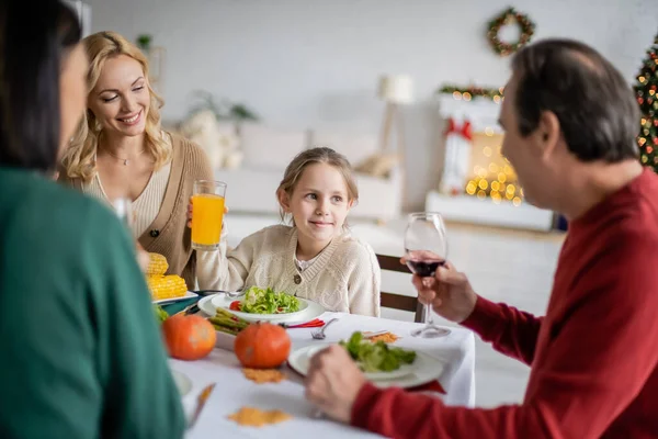 Lächelndes Kind mit Orangensaft blickt verschwommene Großeltern bei Erntedankfeier zu Hause an — Stockfoto