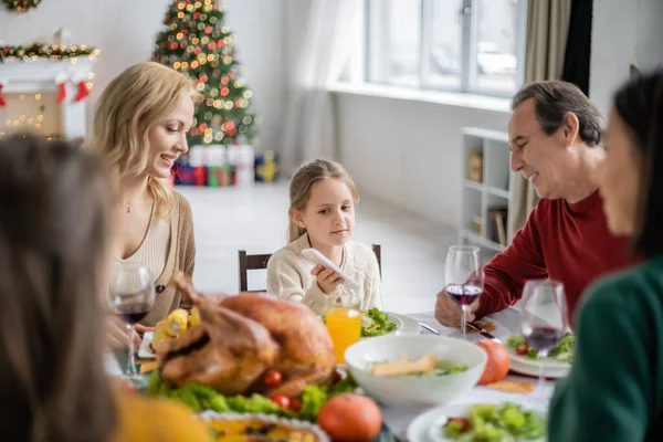 Ребенок держит смартфон рядом с улыбающейся бабушкой и дедушкой во время ужина в честь Дня Благодарения дома — стоковое фото
