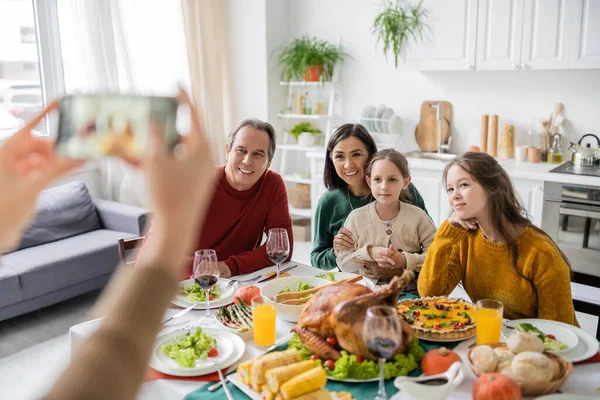 Multiethnic family sitting near thanksgiving dinner while woman taking photo on smartphone at home — Stock Photo