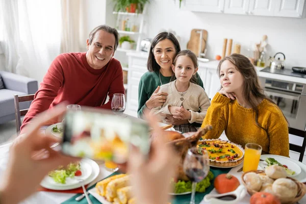 Familia interracial sonriendo cerca de deliciosa cena de acción de gracias y mujer tomando fotos en el teléfono inteligente en casa - foto de stock