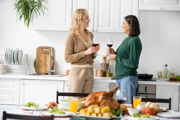Vista lateral de mãe e filha multiétnica positiva segurando vinho e conversando perto do jantar de ação de graças borrado — Fotografia de Stock