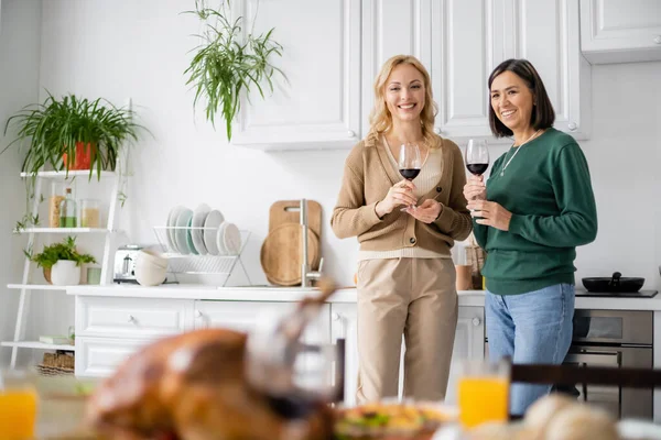 Madre e hija multiétnicas sonrientes sosteniendo copas de vino durante la celebración de Acción de Gracias en casa - foto de stock