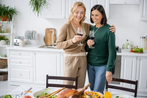 Femme souriante embrassant maman multiethnique avec un verre de vin pendant le dîner de Thanksgiving — Photo de stock