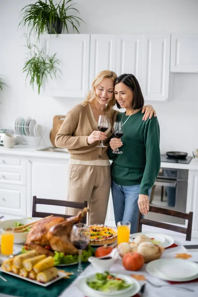 Mujer alegre sosteniendo una copa de vino y abrazando a la mamá multicultural durante el Día de Acción de Gracias en casa - foto de stock