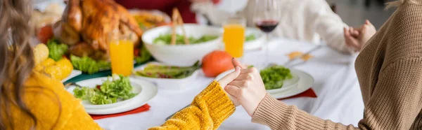 Vista recortada de la familia cogida de la mano cerca de la cena de acción de gracias con pavo en casa, pancarta - foto de stock