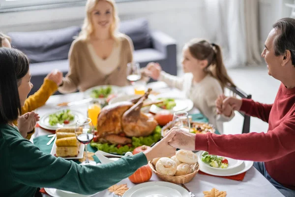 Interracial grandparents holding hands near blurred family and thanksgiving dinner at home — Stock Photo