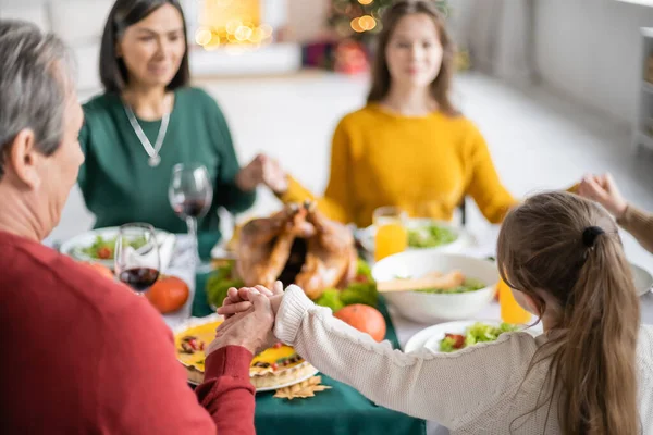 Kid holding hand of grandparent near blurred thanksgiving dinner and family at home — Stock Photo