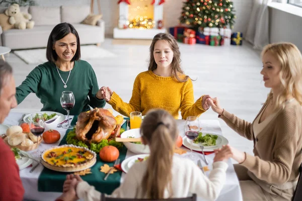 Familia multiétnica sonriente cogida de la mano cerca de pavo de acción de gracias y bebidas en casa - foto de stock