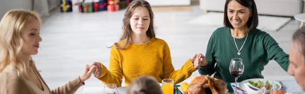 Familia multicultural de la mano cerca de sabrosa cena de acción de gracias en la mesa en casa, pancarta - foto de stock