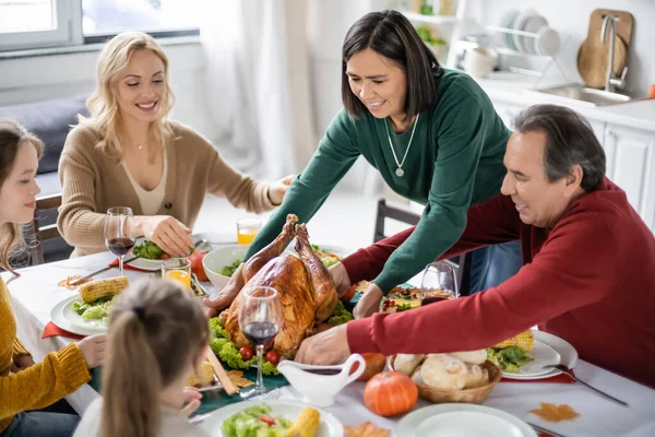 Multicultural woman putting turkey on table near thanksgiving dinner and family at home — Stock Photo