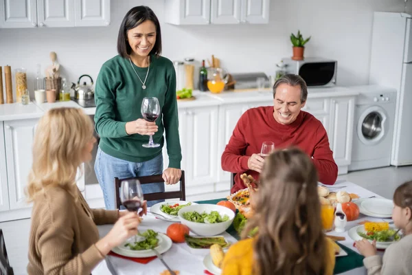 Femme multiethnique tenant un verre de vin près de la famille floue et dîner de Thanksgiving — Photo de stock