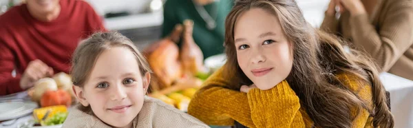 Позитивные девушки смотрят в камеру возле размытой семьи и ужина во время празднования Дня благодарения дома, баннер — стоковое фото