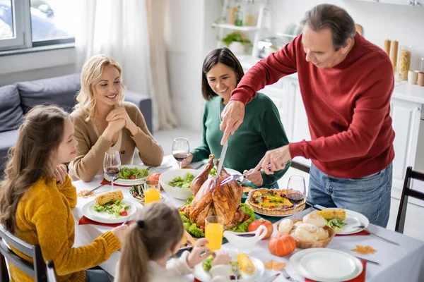 Man cutting turkey near smiling multiethnic family during thanksgiving dinner at home — Stock Photo