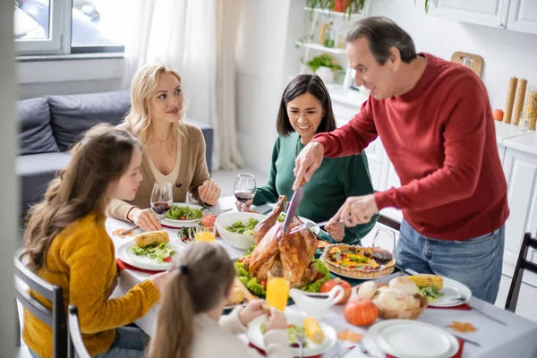 Улыбающаяся женщина смотрит, как родители режут индейку на День Благодарения возле многонациональной семьи дома — стоковое фото