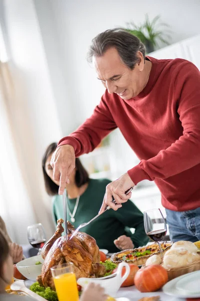 Allegro uomo che taglia tacchino Ringraziamento vicino a cena e la famiglia a casa — Foto stock