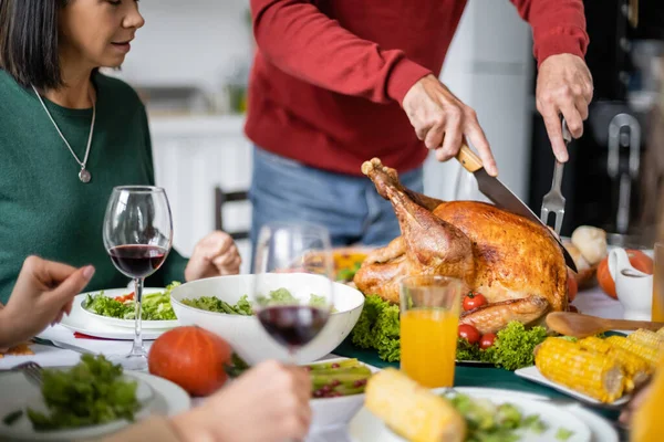 Cropped view of senior man cutting delicious thanksgiving turkey near women and wine at home — Stock Photo