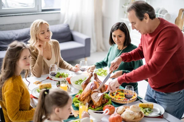 Sonriente familia multiétnica mirando al hombre mayor cortando pavo de acción de gracias en casa - foto de stock