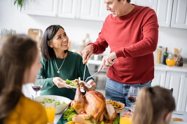 Старший мужчина кладет индейку на тарелку возле межрасовой семьи празднует День благодарения дома — стоковое фото