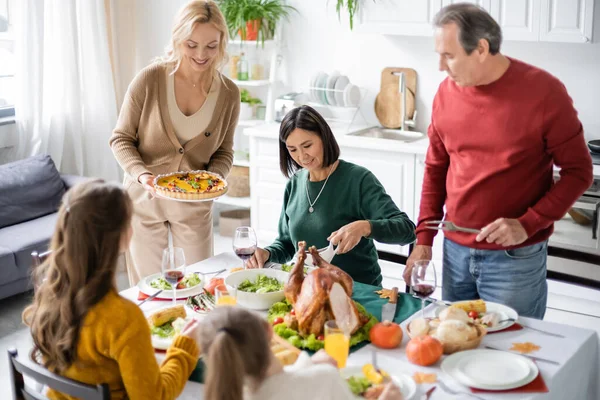 Donna sorridente che tiene deliziosa torta vicino alla famiglia multiculturale e ai bambini durante la cena del Ringraziamento — Foto stock