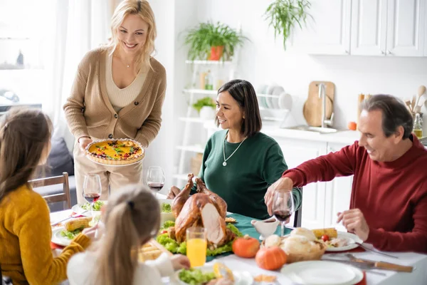 Mujer alegre sosteniendo pastel cerca de padres e hijos multiculturales durante la cena de acción de gracias - foto de stock