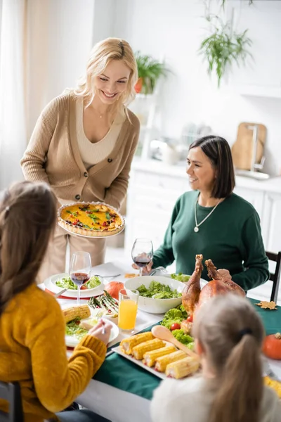 Positive woman holding tasty thanksgiving pie near multiethnic mother and children at home — Stock Photo