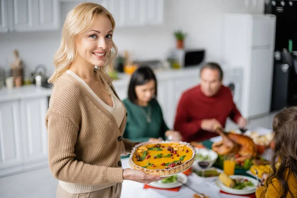 Smiling woman holding thanksgiving pie and looking at camera near blurred family at home — Stock Photo