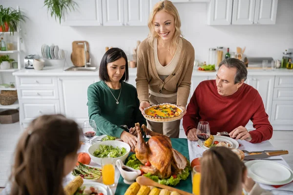 Donna sorridente che tiene torta di ringraziamento vicino ai genitori multiculturali e ai bambini a casa — Foto stock