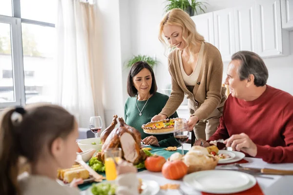 Positive woman holding pie near multiethnic parents and thanksgiving dinner at home — Stock Photo