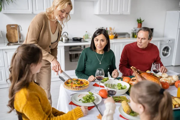 Cheerful woman cutting pie near interracial family and thanksgiving dinner at home — Stock Photo
