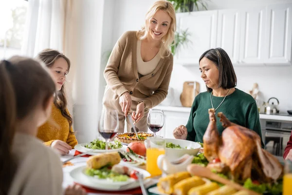 Lächelnde Mutter schneidet Torte neben multikultureller Familie und verschwommener Danksagungs-Truthahn zu Hause — Stockfoto